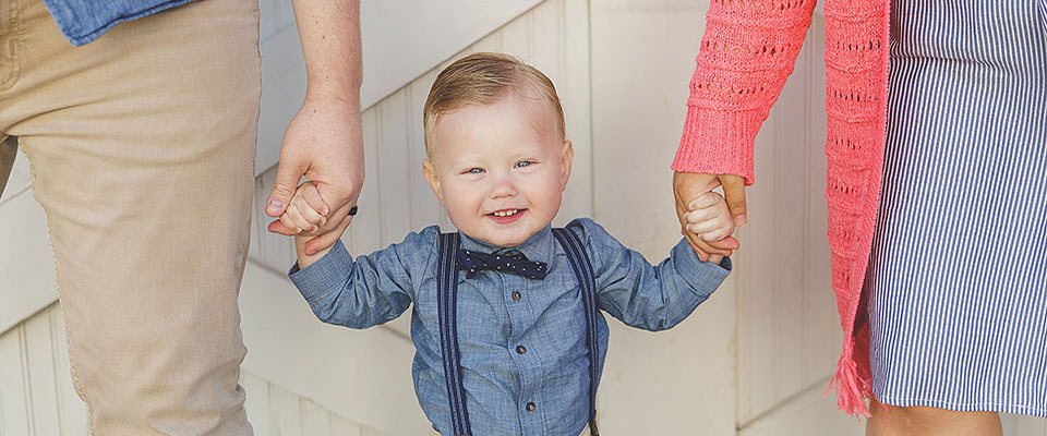 A smiling toddler boy in blue and suspenders holding the hands of his parents