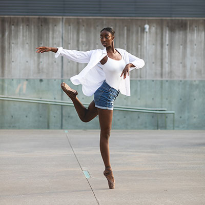 A Black teenage dancer en pointe in a concrete and glass area, wearing a white shirt and denim shorts - dallas dance photography