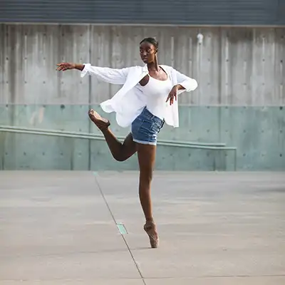 A Black teenage dancer en pointe in a concrete and glass area, wearing a white shirt and denim shorts - dallas dance photography