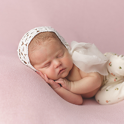 light skinned sleeping newborn baby on pink backdrop, with white bonnet and pants, and hands under her face