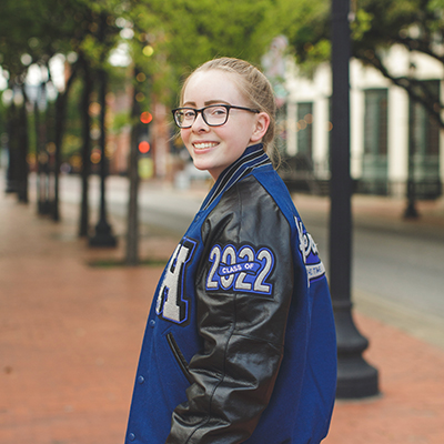 light skinned senior girl in a black and blue letterman jacket turns back to look at the camera - plano senior photographer
