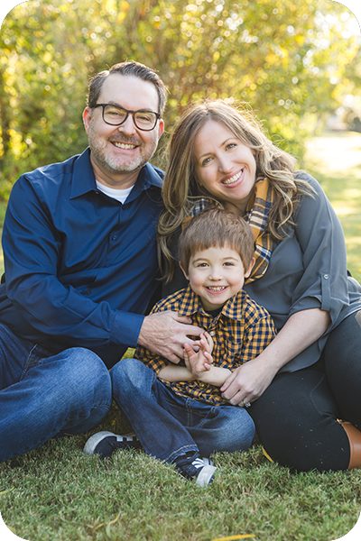 A vertical photo of a family with a dad, mom and young boy sitting on the grass and smiling. They are wearing shades of navy, mustard, gray and black