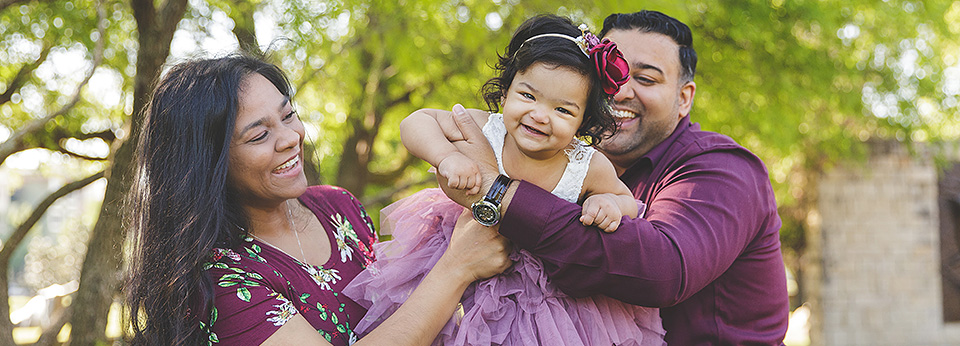 A south Asian family with mom, one year old girl and dad, all wearing shades of maroon and purple.